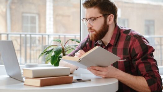 man at table with laptop and stack of books looks through best Bible commentaries