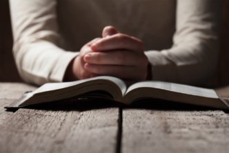 Person's hands folded in prayer behind an open Bible