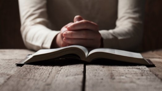 Person's hands folded in prayer behind an open Bible