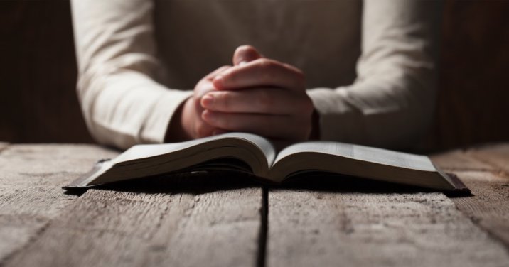 Person's hands folded in prayer behind an open Bible