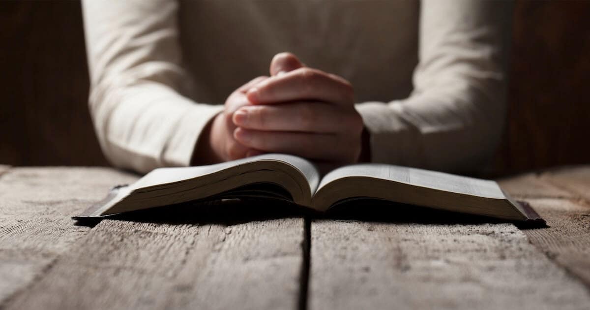 Person's hands folded in prayer behind an open Bible
