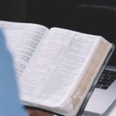 man at desk studies biblical hermeneutics with Bible software on laptop & paper books