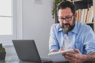 bearded man with a laptop makes notes about Logos 10 feature upgrades in a notebook