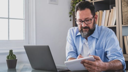 bearded man with a laptop makes notes about Logos 10 feature upgrades in a notebook