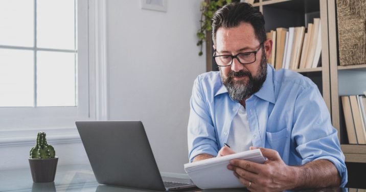 bearded man with a laptop makes notes about Logos 10 feature upgrades in a notebook